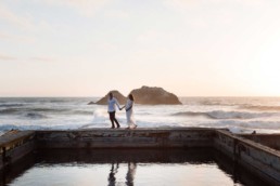 Young Pregnant Woman in Flowing White Summer Dress with the Bay Ocean Waves Crashing at Sunset at Sutro Baths in Lands End Park by San Francisco Family Photographer