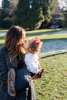 Young Mom Kissing Baby Little Girl Toddler In Red Bow With Sunlight in Volunteer Park by Seattle Family Photographer